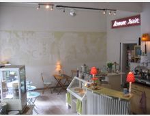 Gemütliches Café mit heller Einrichtung, kleinen Holztischen und Stühlen. Im Vordergrund befinden sich eine Theke mit roten Lampenschirmen, Glasgefäßen und Gebäck, im Hintergrund eine bemalte Wand und ein leuchtendes „komm rein“-Schild.