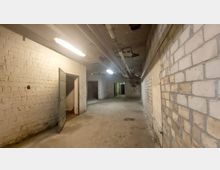 Ein leerer, unterirdischer Korridor mit weißen Ziegelwänden und abgenutztem Betonboden. Die Decke zeigt freiliegende Rohre und Beleuchtungskörper, während geöffnete Türen zu angrenzenden Räumen führen.