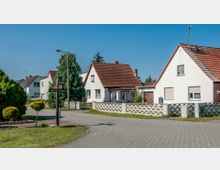 Straßenszene in einem Wohngebiet mit Einfamilienhäusern, die weiße oder helle Fassaden und rote Satteldächer haben. Die Straße ist mit Pflastersteinen ausgelegt, Grünflächen und niedrige Zäune trennen die Grundstücke voneinander.