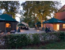 Ein Biergarten mit Schotterboden, umgeben von Bäumen und niedrigen Hecken. Mehrere Holzbänke und Tische mit großen, grünen Sonnenschirmen sind aufgestellt, und ein hölzerner Kiosk mit Verkaufsfenster befindet sich links im Bild.