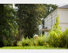 Rechte Seite eines weißen historischen Gebäudes mit Fenstern und einem Balkon mit Geländer, umgeben von einer grünen Gartenlandschaft und hohen Bäumen. Im Vordergrund befinden sich Büsche und Wiesen.