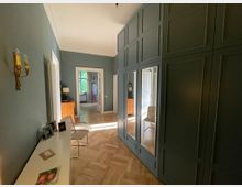 Flur in einer Wohnung mit dunkelgrauen Einbauschränken und integrierten Spiegelflächen an der rechten Wand, einem hellen Holzboden und blau-grauen Wänden. Im Hintergrund führt der Flur zu einem Raum mit großen Fenstern, durch die Tageslicht einfällt.