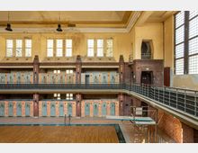 Historisches Hallenbad mit leerem Schwimmbecken, umlaufenden Balkonen aus Metallgeländern und einem Backstein-Interieur. Große Fenster mit Buntglas und Umkleidekabinen mit bunten Türen prägen den Raum.