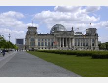Der Reichstag in Berlin mit seiner markanten Glaskuppel, flankiert von historischen Gebäudeflügeln und beflaggt mit deutschen Flaggen. Im Vordergrund befinden sich eine breite Promenade und Grünflächen, im Hintergrund der Fernsehturm und moderne Gebäude.