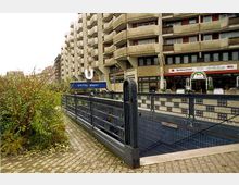 Ein U-Bahn-Eingang mit blauer Beschilderung „Spittelmarkt“ vor einem Wohn- und Geschäftsgebäude mit Balkonen und Läden im Erdgeschoss; im Vordergrund ist eine gepflasterte Fläche mit Begrünung sichtbar.