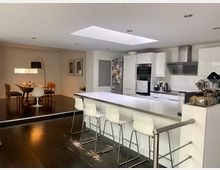 Moderne Küche mit weißem Interieur, einer großen Kücheninsel und vier Barhockern. Im Hintergrund befindet sich ein Essbereich mit Holztisch, verschiedenen Stühlen und einer Stehlampe; Holzfußboden und ein großes Oberlicht prägen den Raum.