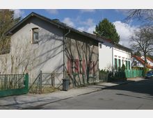 Straßenszene mit Wohnhäusern, darunter ein graues Gebäude mit roten Fensterläden und ein weißes Haus mit türkisfarbenen Fensterläden. Vor den Häusern verlaufen Zäune und ein Gehsteig, im Hintergrund sind Bäume und ein Auto zu sehen.