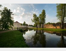 Parklandschaft mit einem Schloss im Hintergrund, flankiert von einer historischen Mauer und großen Bäumen am Ufer eines Wassergrabens, der die Umgebung spiegelt. Eine kleine Holzbrücke führt über das Wasser, eingebettet in gepflegte Wiesen.