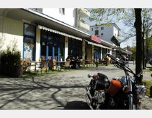 Straßencafé mit blauen Fenstern und einer Markise, davor mehrere Tische mit Stühlen auf einer kleinen Terrasse; ein Motorrad steht im Vordergrund neben Bäumen und einem Gehweg. Im Hintergrund sind Wohn- und Geschäftsgebäude zu sehen.