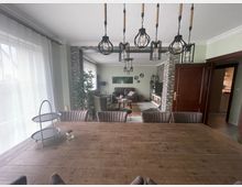 Ein modernes Wohnzimmer und Esszimmer in offener Raumgestaltung. Im Vordergrund ein großer Esstisch aus Holz mit gepolsterten Stühlen und einer Hängelampe mit mehreren Glühbirnen in Käfigdesign, im Hintergrund ein Wohnzimmer mit einem Sofa, einem Fernseher und einer grauen Steinwand.