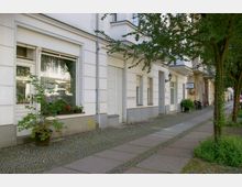 Straßenszene in einer Wohngegend mit einem weiß gestrichenen Mehrfamilienhaus, großen Fenstern mit Blumenkästen und einer gepflasterten Gehwegfläche mit Bäumen am Straßenrand.