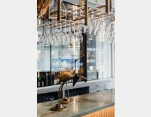 Theke einer modernen Bar mit hängenden Weingläsern an einem goldfarbenen Glasgestell und goldenen Vogel-Skulpturen auf der Arbeitsfläche; im Hintergrund sind Weinschränke mit Flaschen erkennbar.
