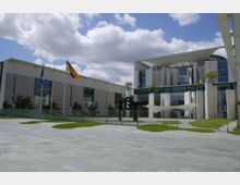 Moderne Regierungsgebäude mit heller Fassade, großen Glasfenstern und Säulen, davor ein umzäunter Hof mit gepflegtem Rasen. Im Vordergrund wehen die deutsche Flagge und die Europaflagge, der Himmel ist leicht bewölkt.