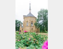 Historische Kirche mit hellem Backstein und neugotischen Elementen, darunter spitzbogige Fenster und ein hoher Turm mit einer Spitze. Im Vordergrund blühende Büsche, umgeben von Bäumen und einer Wiesenlandschaft.