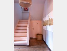 Treppenhaus eines Wohnhauses mit pastellrosa Treppe und hölzernem Boden. Rechts an der Wand befinden sich zwei Bilder mit Skyline-Motiven, darunter ein geflochtener Korb, und eine kleine Tür unter der Treppe.