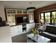 Wohnzimmer mit Holz- und Landhausstil-Elementen. Ein weißes Regal mit Glastüren und Schubladen integriert einen Fernseher, daneben große Holzfenster mit grünen Rahmen und Pflanzen; im Vordergrund ein Couchtisch mit Blumen und eine dunkle Ledercouch.