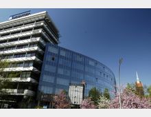 Moderne Bürogebäude mit Glasfassaden und abgerundeten Ecken, neben einem älteren Kirchturm mit rotem Ziegelgebäude. Im Vordergrund blühende Bäume und eine Straßenlaterne, bei klarem blauen Himmel.