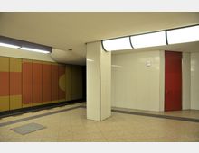 Untergrundhalle mit beigen Bodenfliesen und weißen Wandfliesen, einer roten Tür rechts und einer Wand auf der linken Seite mit farbigem geometrischem Muster in Gelb und Rot. Die Decke ist beige mit moderner Beleuchtung, und ein quadratischer Pfeiler teilt den Raum.