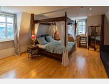 Schlafzimmer mit einem dunklen Himmelbett, umgeben von weißen Vorhängen, auf hellem Holzfußboden. Im Raum befinden sich zwei Fenster mit Vorhängen, ein Spiegel, eine Kommode und rote Nachttischlampen.