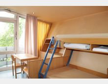 Kleines, schlichtes Zimmer mit Etagenbett aus Holz, blauer Metallleiter, Tisch und zwei Stühlen. Links befindet sich ein Fenster mit halbtransparenten weißen und gelben Vorhängen, das Tageslicht hereinlässt.