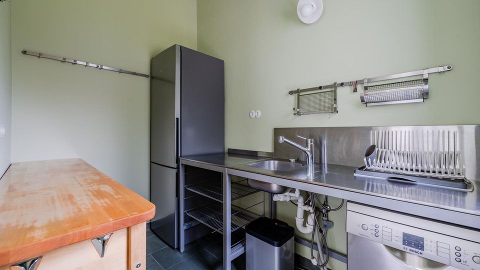 Schlichte Küchenzeile mit grünen Wänden, Edelstahl-Arbeitsplatte und Spülbecken, daneben ein hoher schwarzer Kühlschrank. Unter der Arbeitsplatte stehen eine Spülmaschine und offene Regalböden; an der Wand sind Abtropfgestell und Aufhängeschienen montiert, links ein kleiner Holzarbeitstisch.