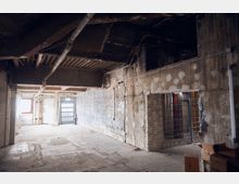 Ein leerer, industrieller Raum mit rohem Beton, freiliegenden Rohren an der Decke und abgenutzten Wänden. Im Hintergrund befindet sich eine Metalltür mit einem kleinen Fenster, rechts sind Gitter und offene Kabel sichtbar.