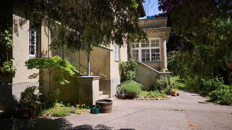 Seitliche Ansicht eines hell verputzten Wohnhauses mit Treppe und kleinem Eingangsbereich mit Säule und großen Fenstern. Ein gepflasterter Hofweg führt durch dichten, schattigen Gartenbewuchs mit Sträuchern, Topfpflanzen und Gießkanne am Rand.