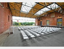 Open-Air-Veranstaltungsbereich mit Reihen von weißen Stühlen auf einem gepflasterten Boden, umgeben von roten Backsteinwänden einer ehemaligen Industriehalle. Das Dach besteht aus Metall- und Glasstrukturen, die teilweise geöffnet sind, und im Hintergrund befindet sich eine Grünfläche mit Blick ins Freie.
