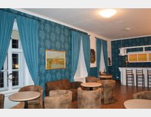 Ein Innenraum mit blauen, gemusterten Wänden und bodenlangen Vorhängen vor weißen Fenstern. Der Raum ist eingerichtet mit braunen Sesseln, runden Tischen und einer weißen Bar mit Holzstühlen.