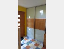 Eingangsbereich mit einer hölzernen Tür, gelben Wänden und mehrfarbig gefliesten Boden. Rechts befinden sich Schiebetüren mit Milchglas und einem Abschnitt aus perforiertem Holzdekor.