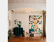 Ein stilvoll eingerichtetes Atelier mit hohen Decken und dekorativen Stuckverzierungen. Der Raum enthält abstrakte Gemälde an den Wänden, eine grüne Designer-Kommode, Pflanzen und eine Arbeitsfläche mit Metallgestell und Hockern auf einem Holzboden.