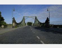 Grüne Stahlbrücke mit geschwungenen Bögen und dekorativen Statuen an den Seiten, darunter eine gepflasterte Fahrbahn mit jeweils einer Autospur pro Richtung. Die Brücke ist von Laternen und Bäumen im Hintergrund eingerahmt, unter klarem Himmel.