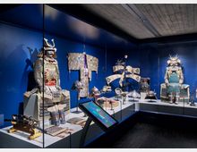 Ausstellungsraum eines Museums mit einer Glasvitrine, die traditionelle japanische Samurai-Rüstungen, Helme und Schwerter zeigt. Im Hintergrund hängen zeremonielle Gewänder und Fächer vor einer blauen Wand, während im Vordergrund ein interaktives Display positioniert ist.