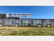 Eine Betonwand mit bemalten Landschafts-Motiven, darunter Bäume, Wiesen und Gebäude, vor einer grasbewachsenen Fläche. Darüber befindet sich ein großes Werbeschild für "Tiefbau- und Schüttguthandel" mit Kontaktdaten und der Ortsangabe "Schwedt".