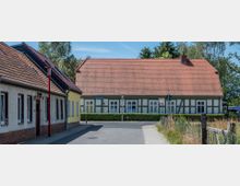 Fachwerkhaus in einer ländlichen Umgebung mit rotem Ziegeldach und umliegenden Gebäuden, einer Straße sowie Gehwegen im Vordergrund. Im Hintergrund sind Bäume und ein heller Himmel zu sehen.