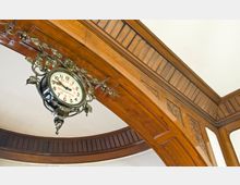 Eine antike Wanduhr mit dekorativem Metallrahmen und floralen Mustern hängt unter einem reich verzierten hölzernen Bogen, der mit Schnitzereien und Holzverkleidungen gestaltet ist.