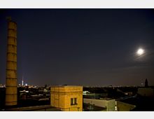 Nachtsicht einer Stadt mit beleuchtetem Himmel, einer großen Fabrikschornstein-Silhouette links und einem gelben Backsteingebäude im Vordergrund. Im Hintergrund ist der Fernsehturm und eine hell leuchtende Vollmondszene sichtbar.