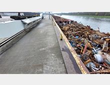 Ein Schiffsanlegeplatz mit einem Frachtschiff, das mit Metallschrott beladen ist. Links entlang der Betonkai sind Schienen sichtbar, und rechts befindet sich ein Fluss mit bewaldetem Ufer im Hintergrund.