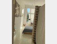 Helles Schlafzimmer mit schlichtem Design: Ein niedriges Bett mit blauer Bettwäsche steht auf einem weiß glänzenden Boden. An der linken Wand hängen minimalistische Kunstwerke, während große Fenster mit halbtransparenten Vorhängen den Raum mit Tageslicht erhellen. Ein Stapel Pappkartons und ein Kleiderständer befinden sich im Hintergrund.