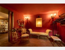 Edler Raum in einem Barockstil mit roten Wänden, goldverzierten Möbeln und Holzboden. Zu sehen sind ein gepolsterter Stuhl, eine goldene Konsole mit Spiegel, ein dekorativer Kerzenleuchter in einer Wandnische sowie eine Liege und Kleiderhaken an der Wand.