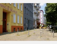 Straßenansicht einer urbanen Gasse mit mehrstöckigen Wohnhäusern in verschiedenen Farben (Gelb, Grau, Weiß und Rosa). Der gepflasterte Weg ist von Pollern gesäumt, während auf der rechten Seite Bäume, Fahrräder und Außensitzbereiche von Cafés sichtbar sind.