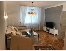 Wohnzimmer mit Parkettboden, einer hellen Eckcouch, einem Glastisch und einem Teppich. Links steht eine Wandlampe, in der Mitte ein Kronleuchter, und gegenüber der Tür ein Fernseher auf einem weißen Schrank; Vorhänge bedecken das Fenster.