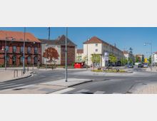 Eine städtische Straßenkreuzung mit einem kleinen Kreisverkehr, umgeben von Gebäuden mit roten Ziegeldächern und heller Fassade. Eine Hauswand zeigt ein großes Wandbild eines Baumes, im Vordergrund blühen gelbe Blumen im Kreisverkehr, und vereinzelte Fußgänger und Autos sind sichtbar.