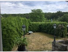 Ein kleiner Garten mit einer Rasenfläche, umgeben von hohen immergrünen Hecken. Im Vordergrund stehen zwei schwarze Laternen und ein großer Pflanzentopf; im Hintergrund ist ein einzelner Baum sowie ein Wald unter bewölktem Himmel zu sehen.