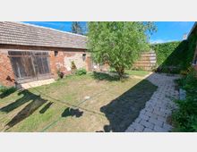 Hinterhof mit einer offenen Rasenfläche, einem Baum in der Mitte und gepflasterten Wegen. Im Hintergrund sind eine Backsteinmauer mit Holztor, eine Holzwand sowie eine mit Efeu bewachsene Wand zu sehen.