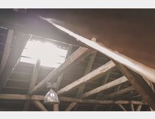 Holzgebälk einer Dachkonstruktion mit sichtbaren Querbalken und einer großen Dachöffnung, durch die helles Tageslicht einfällt. Eine einzelne Metalllampe hängt von einem der Balken.