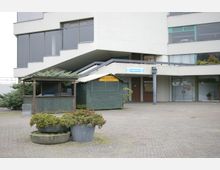 Außenbereich eines modernen Gebäudes mit großen Glasfenstern und Betonfassade. Im Vordergrund stehen bepflanzte Kübel und ein kleines, grünes Holzhäuschen mit der Aufschrift „Festzentrum Trabrennbahn“.