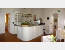 Heller Gastraum mit einer weißen Holztheke, die mit Geschirr und dekorativen Silberkannen ausgestattet ist. Der Raum hat Parkettboden, offene weiße Doppeltüren führen zu weiteren, gemütlich eingerichteten Räumen, und Tische mit weißem Dekor stehen bereit.