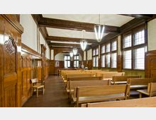 Ein historischer Saal mit Holztäfelungen an den Wänden, Reihen aus hölzernen Sitzbänken und großen Fenstern mit kleineingeteilten Scheiben. Deckenleuchten und geschnitzte Verzierungen betonen den klassischen Stil der Einrichtung.