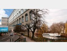 Eine Straßenansicht mit einem großen Ziegelgebäude auf der linken Seite, das viele hohe Fenster und ein Schild mit der Aufschrift „cine+“ hat. Rechts führt ein Weg an einem Fluss entlang, auf dem ein weißes Ausflugsschiff liegt, umgeben von kahlen Bäumen.